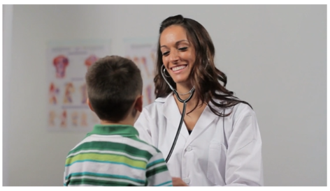 A white, female clinician in a white coat uses a stethoscope on a young boy in a green striped shirt.