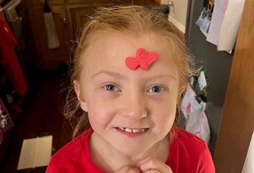 Londyn, a Children's Mercy patient, smiles for the camera with a piece of red clay stuck to her forehead