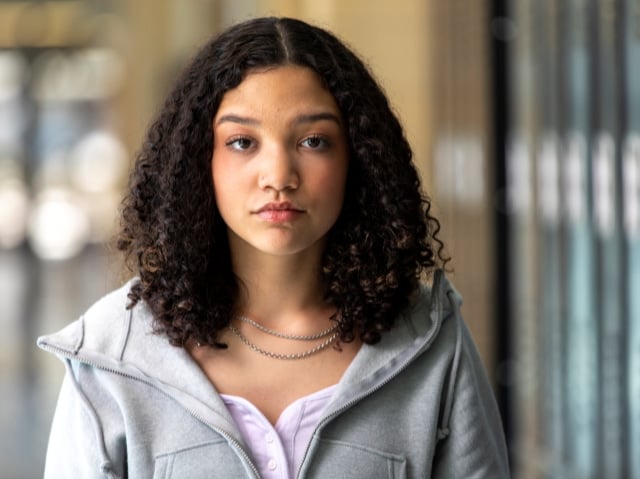 A teen girl with curly brown hair in a gray hoodie looks directly at the camera