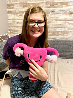 Taylor Stewart smiling and holding a pink pillow in the shape of a uterus with fallopian tubes and ovaries. The pillow has a smiley face.