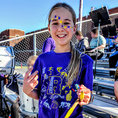 A young teen stands in the bleachers of a stadium surrounded by various drums. She is wearing a purple and yellow shirt with her school name and logo and holds yellow drumsticks. She has purple and yellow facepaint dots on her forehead and cheeks.