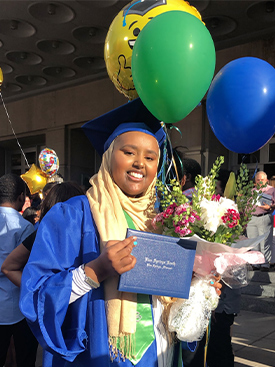 Munira Nuru smiling while wearing a graduation cap and gown, and holding her diploma, flowers and balloons.