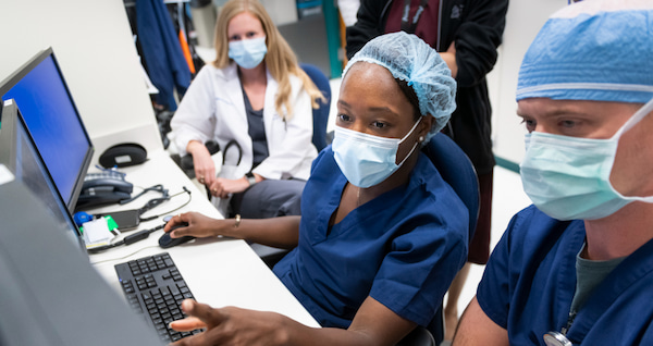 A masked, Black woman wearing looks at a computer monitor as two of her colleagues look on.