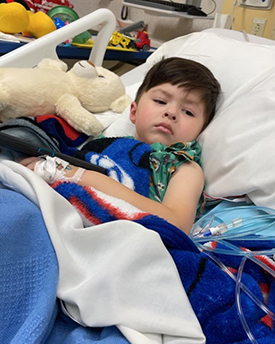 Elijah after surgery at Children's Mercy. He is laying in a hospital bed with a teddy bear.