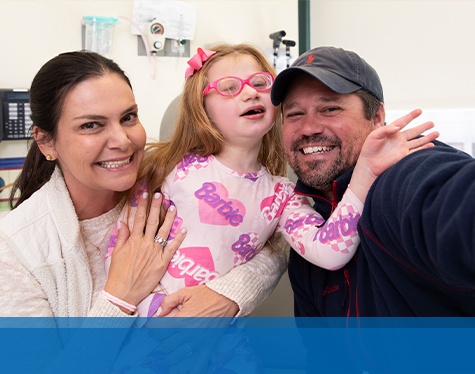 A young girl wearing a pink boy and eyeglasses smiling in between her mother and father at Children's Mercy.