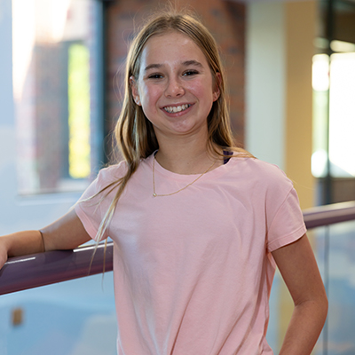 Portrait of a smiling girl with long blond hair in a pink tee shirt, photographed indoors with a blurred background.