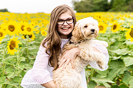 Taylor Stewart standing and smiling in a field of sunflowers while holding a dog. She is wearing a white dress and eyeglasses.
