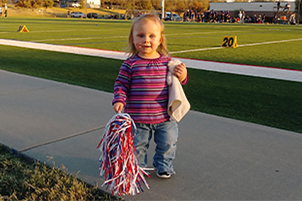 Mercy White stands near a football field smiling with a pom pom in one hand and a stuffed animal/blanket in the other.