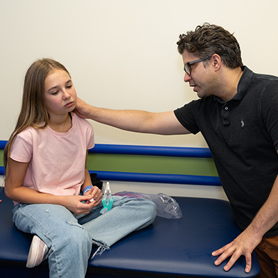 A man with curly dark hair and glasses, wearing a black polo shirt, reaches toward a girl sitting on a clinic exam table. He is touching her cheek as part of a medical examination. The girl is a young teen with long blonde hair. She is wearing a pink shirt and jeans.