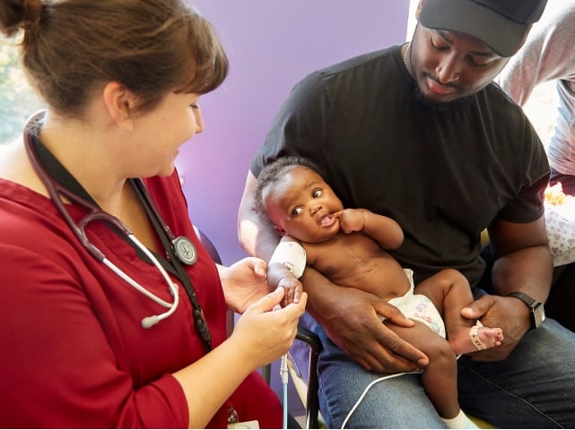 A health care provider in a red shirt with a stethoscope examines a Black baby girl wearing a diaper and cuff on her arm. She is held by a Black man in a black shirt and baseball cap.