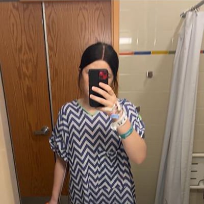 A teen takes a hospital bathroom selfie, her face obscured by her cell phone. She’s wearing a blue and white hospital gown, and we can see a sink in the foreground and a shower curtain in the back. 