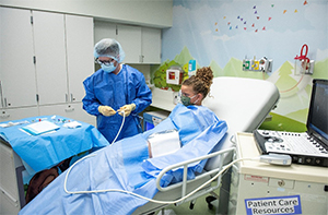 A A Children's Mercy nurse wearing a blue gown, gloves, and a hat and a young patient wearing a face mask and covered with a blue sheet.