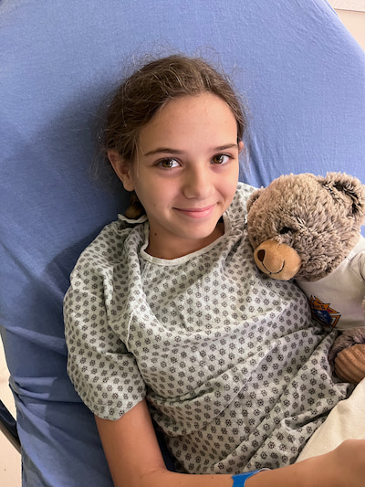A young girl in a hospital gown lies in a bed hugging a teddy bear