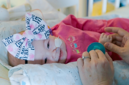 A smiling baby girl with a large bow on her head and a tube leading to her nose lays on her side and smiles. An adult holds her hand and a blue pacifier.