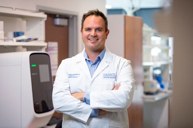 Dr. Craig Smail stands in his lab with his arms crossed and he's smiling at the camera. He wears a white lab coat.