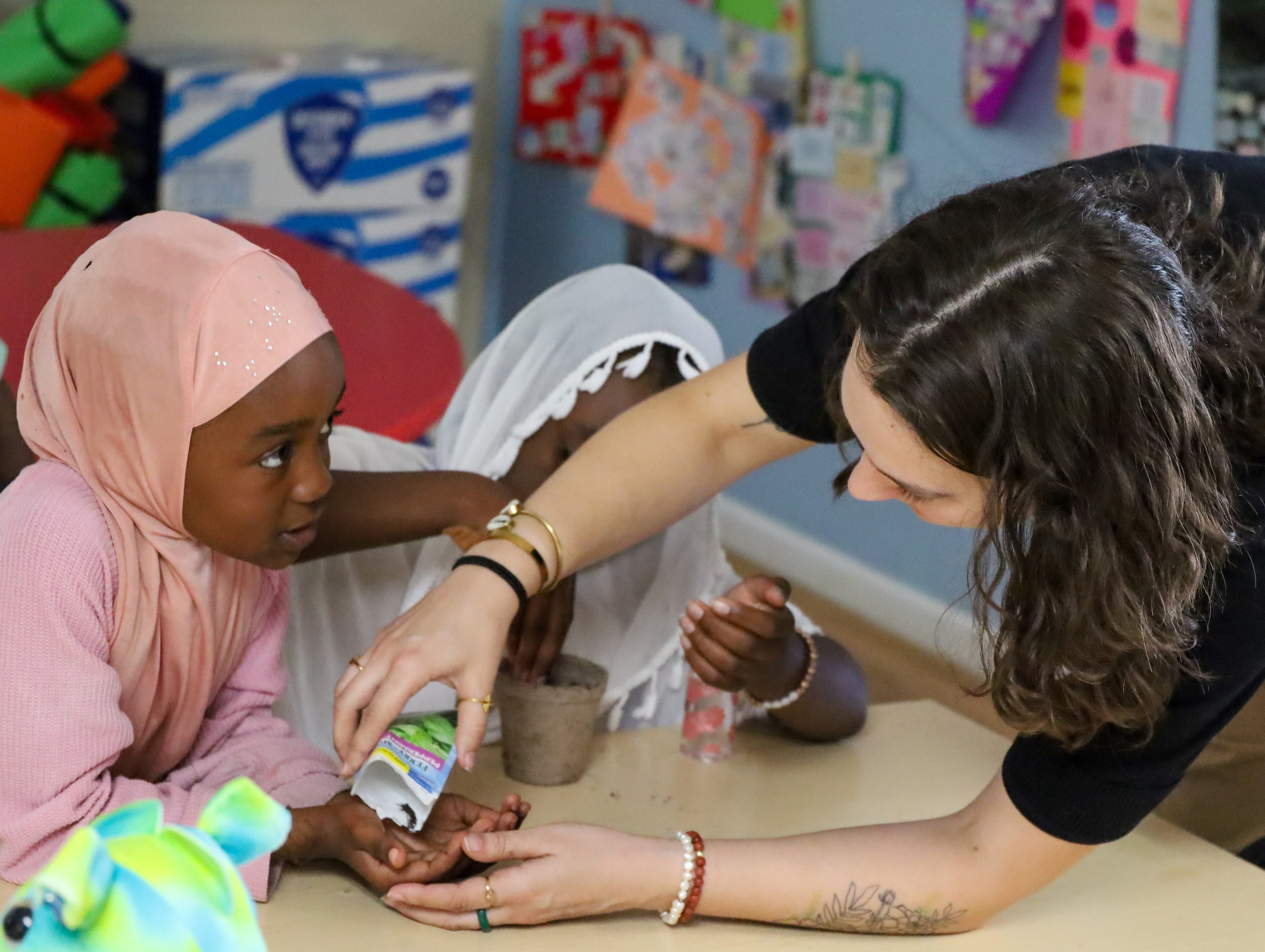 An adult helps two children with a hands-on activity at a classroom table.