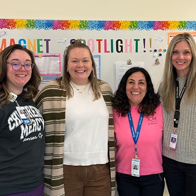 Four women stand in front of a colorful bulletin board. Three are wearing visible hospital badges and all are smiling. The woman on the left is wearing a sweatshirt that reads “Children’s Mercy.”  