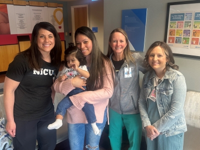 A baby and his mother pose with three hospital team members in a hospital waiting room. One woman has a t-shirt with “NICU” on it, and all three are wearing hospital name badges.