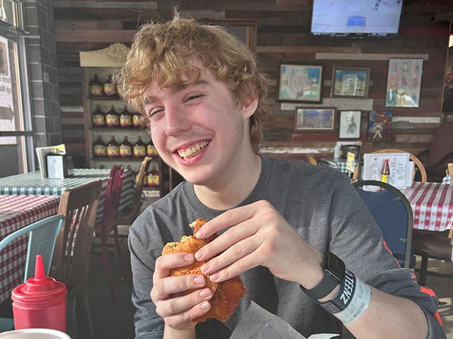 A teenage boy with blond hair sits at a restaurant table holding a chicken sandwich, with a food basket and drink on the table.