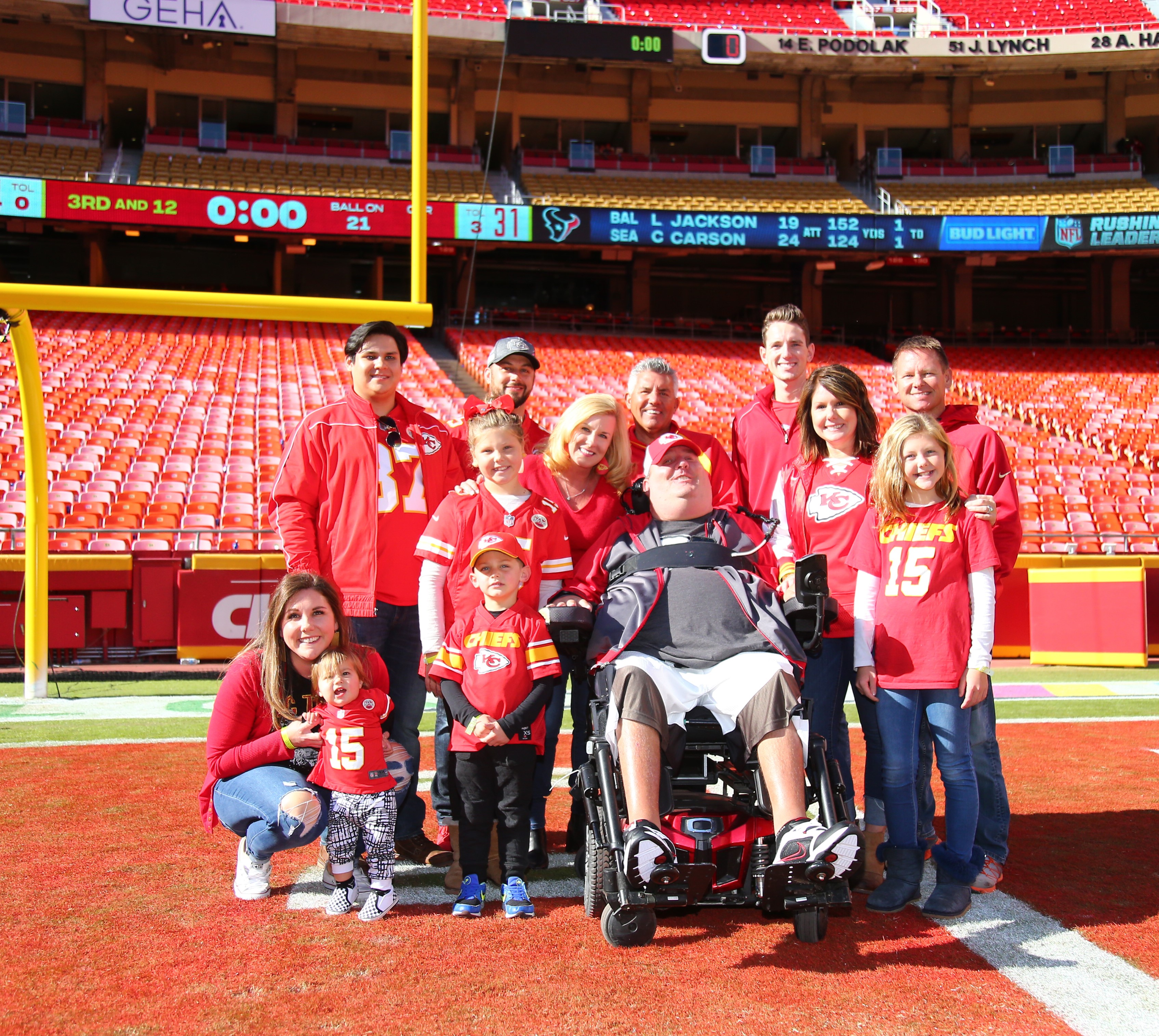 The Baier family (13 people) on the football field at Arrowhead Stadium. They are smiling and wearing red Kansas City Chiefs shirts.