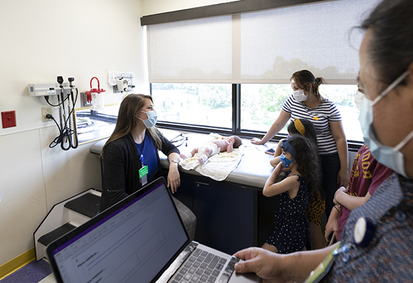 a man who is a language coach stands and holds his laptop computer with a form displayed on the screen while looking at a female physician speak to a mom who is standing next to her two little girls, while mom’s infant lays on an examination table during a clinic visit.