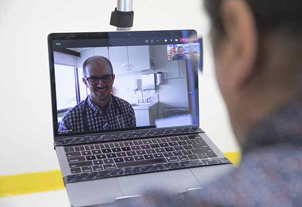 a CM physician’s face is on a laptop computer screen smiling and a Cultural Language Coaching Program male staff member is also on the laptop computer screen in a small box in the top right-hand corner.  Both are virtually meeting with a man who is holding the laptop computer, with only the back of his head shown.