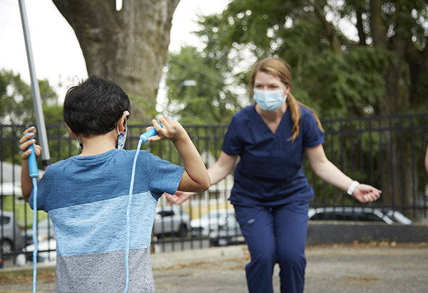 a female dressed in clinical navy-blue scrubs  with mid-length sandy brown hair is holding her hands outstretched pretending to hold an invisible jump rope, while a little boy holds a blue jump rope in his hands, with the jump rope facing his back.