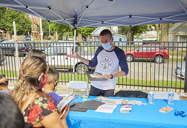 a male in a mostly white t-shirt w/navy blue mid-length sleeves with a CM logo on the front, is holding a black folder and colorful kiddie stickers in one hand.  There are 5 people lined up at his table where he is handing out pamphlets and information on healthcare