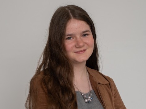 A teenage girl with wavy brown hair is wearing a gray shirt and a brown jacket.
