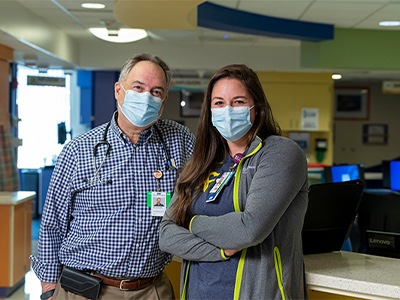 Dr. Alan Gamis with nurse, Shauna Beckett. They are inside Children's Mercy hospital with face masks on, looking at the camera.