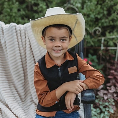 A bright-eyed five-year-old boy sits on a garden bench. He’s smiling and wearing a cowboy hat, western-style shirt and blue jeans.