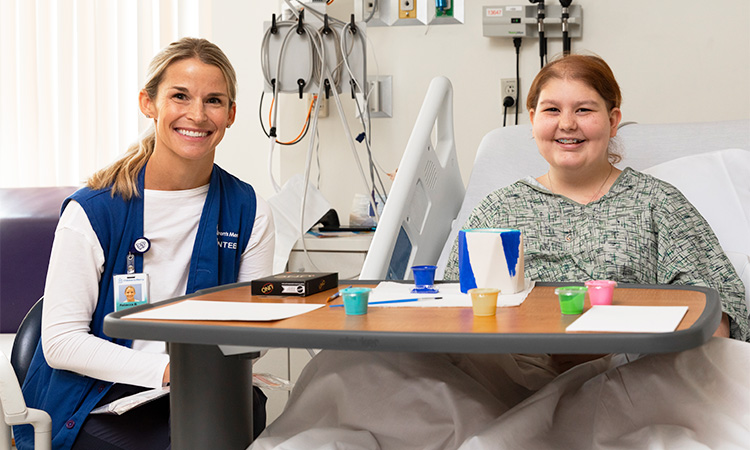 Children's Mercy adult volunteer smiling while sitting next to a patient who is smiling in a hospital bed.