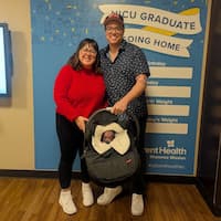 Mom and dad holding baby carrier smiling in front of NICU graduate banner.