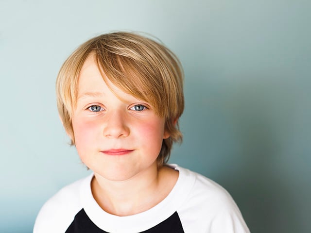 A grade school or middle school aged white boy with blond hair and blue eyes looks directly at the camera with a slight smile.