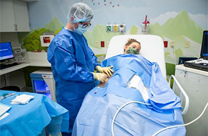 A A Children's Mercy nurse wearing a blue gown, gloves, and a hat and a young patient wearing a face mask and covered with a blue sheet. The nurse is giving the patient numbing medicine through the arm.