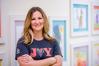 Amy Findlay smiling and wearing a Children's Mercy shirt that reads, "JOY" with the "o" being a heart shape. She is at Children's Mercy and standing in front of multiple artwork done by CM patients.
