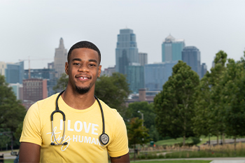 Ijon Howard is smiling and standing outside. The Kansas City, Mo. skyline is behind him and he is wearing a stethoscope around his neck and a shirt that reads, "I love tiny humans."