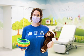 Lenore Flemming is inside a Children's Mercy patient room. She is wearing a face mask and holding a bandaged teddy bear in one hand and a toy in her other hand.