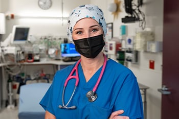 Ugne Krupskis is standing in an exam room at Children's Mercy. She is wearing a scrub top, a scrub cap, a face mask and a stethoscope around her neck.