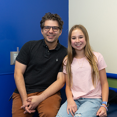 A man with curly dark hair and glasses is wearing a black polo shirt and brown pants. He is sitting on the edge of a clinic exam table with a young teen girl. She has light skin and long blond hair and wears a pink tee shirt and jeans. Both are smiling at the camera.