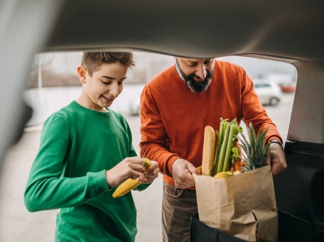 A man in an orange sweater and a boy in a green sweater unloading groceries from a car trunk. A paper bag filled with fresh produce, including celery, pineapple, and a baguette, is visible, while the child holds a banana.