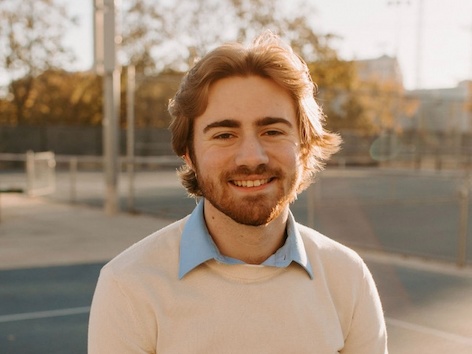 A young adult man with a beard smiles in an outdoor setting.