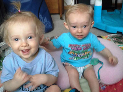The Barker twins, McKinzie and Hudson, sitting on a blanket on the floor and smiling.