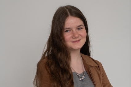 A teenage girl with long, wavy hair is crossing her arms and wearing a gray shirt and a brown jacket.