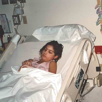 A young patient lies in a hospital bed, covered with blankets, in a clinical room.