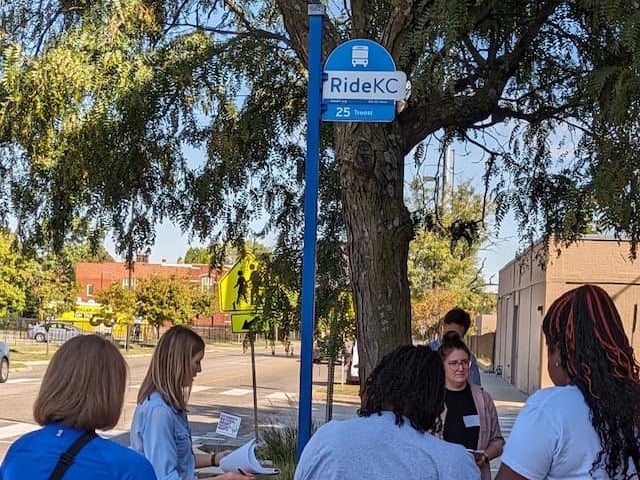 Group of people gathered at a RideKC bus stop during a community assessment training, standing on a sidewalk beneath a tree and reviewing materials near a marked bus stop sign.