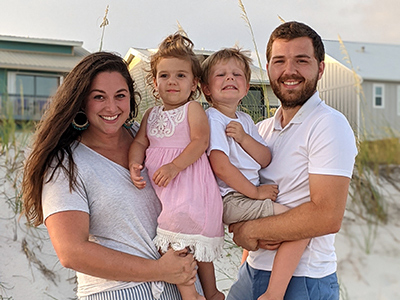Shauna Beckett holder her daughter, Hadley, while standing next to her husband, Luke, who is holding their son, Corbin. They are on the beach and smiling.