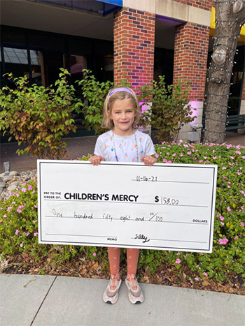 A young girl named Tilly smiling and holding a big check made out to Children’s Mercy for one hundred fifty-eight dollars, dated November 16, 2021.
