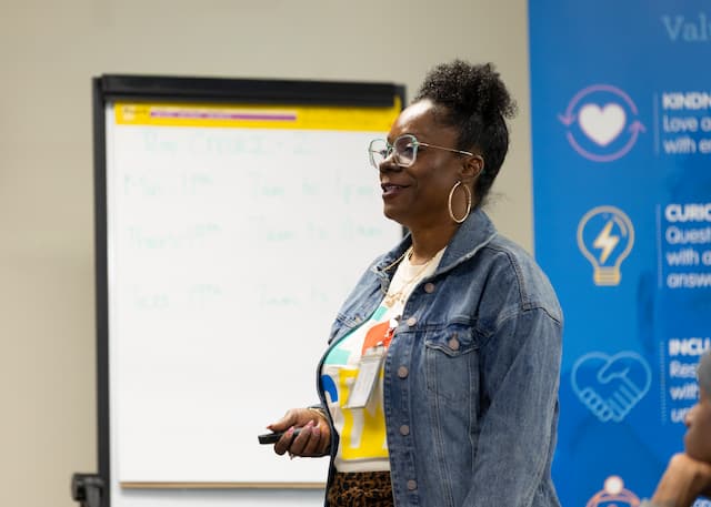Person standing at the front of a meeting room giving a presentation, wearing glasses and a denim jacket, with a whiteboard and informational banner in the background.