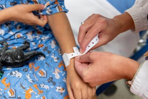 Child arm in blue hospital gown resting on child lap with nurses hands clipping patient wrist band on child's wrist. 